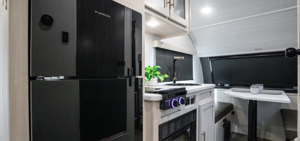 Compact travel trailer interior with refrigerator, kitchenette featuring stove and sink, overhead cabinets, and a rear dinette beneath blackout window shades.