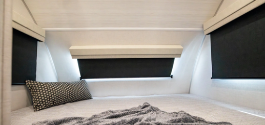 Compact RV sleeping area with a mattress, accent pillow, blanket, and blackout window shades on three sides.