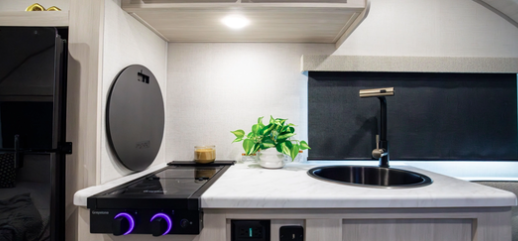 Compact RV kitchenette with a cooktop featuring illuminated knobs, round sink, black faucet, overhead cabinet, and small decorative plant.