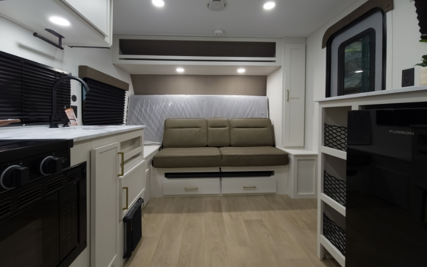 Compact travel trailer interior with kitchenette, storage shelving, rear sofa seating, overhead cabinets, and a fold-down bed stored behind the couch.