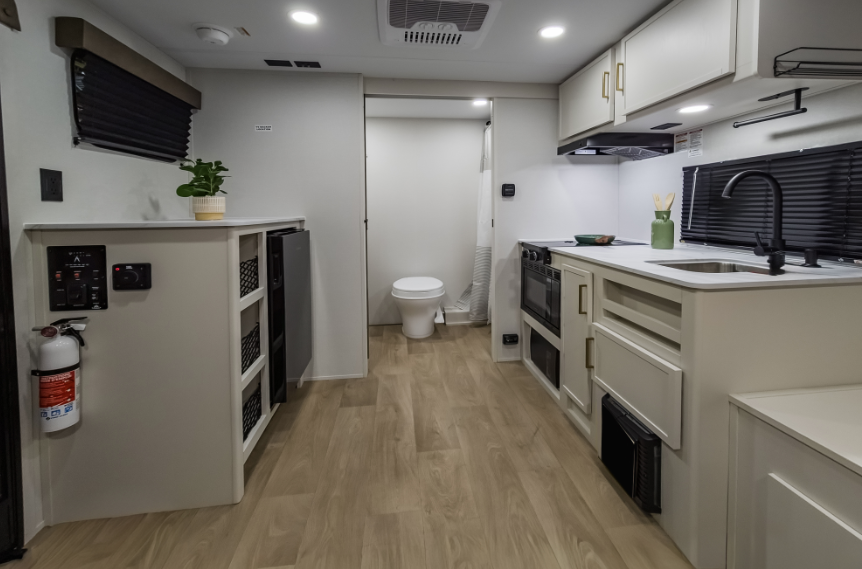 Compact travel trailer interior with kitchenette, sink, stove and oven, storage shelving, and rear bathroom visible through the hallway.