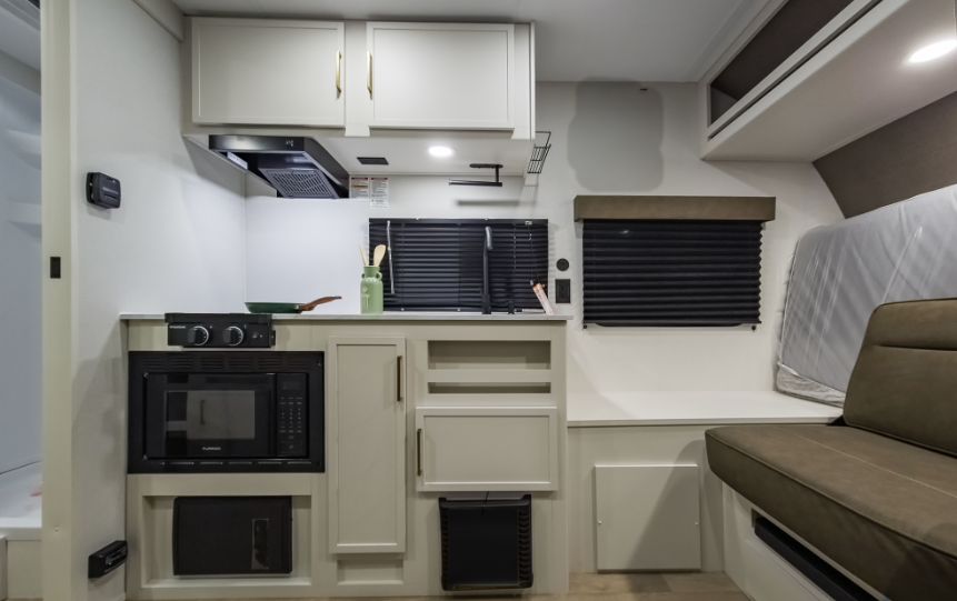 Compact RV kitchenette with microwave, cooktop, sink, overhead cabinets, blackout window shades, and sofa seating beside a fold-down mattress.