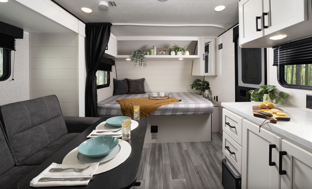 Compact travel trailer interior with dinette seating, kitchenette, overhead storage, and rear bed decorated with pillows and plants.