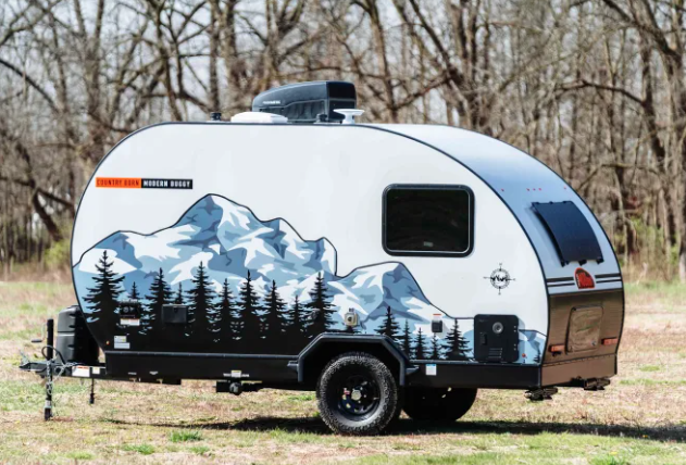 Compact teardrop-style travel trailer with mountain and forest graphics parked outdoors on grass near trees.