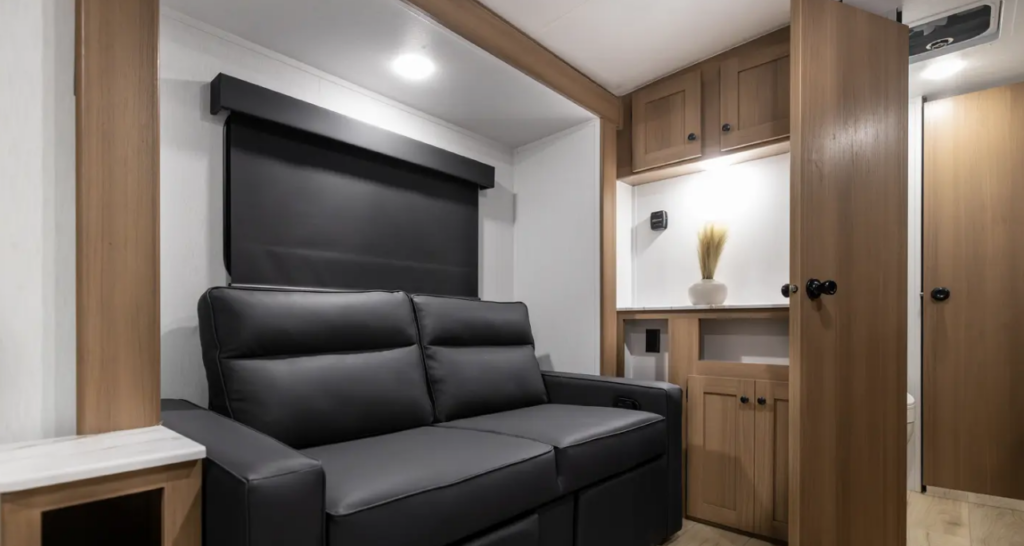 RV interior with a black leather loveseat, blackout window shade, wood cabinetry, and hallway leading toward the bathroom and bedroom.