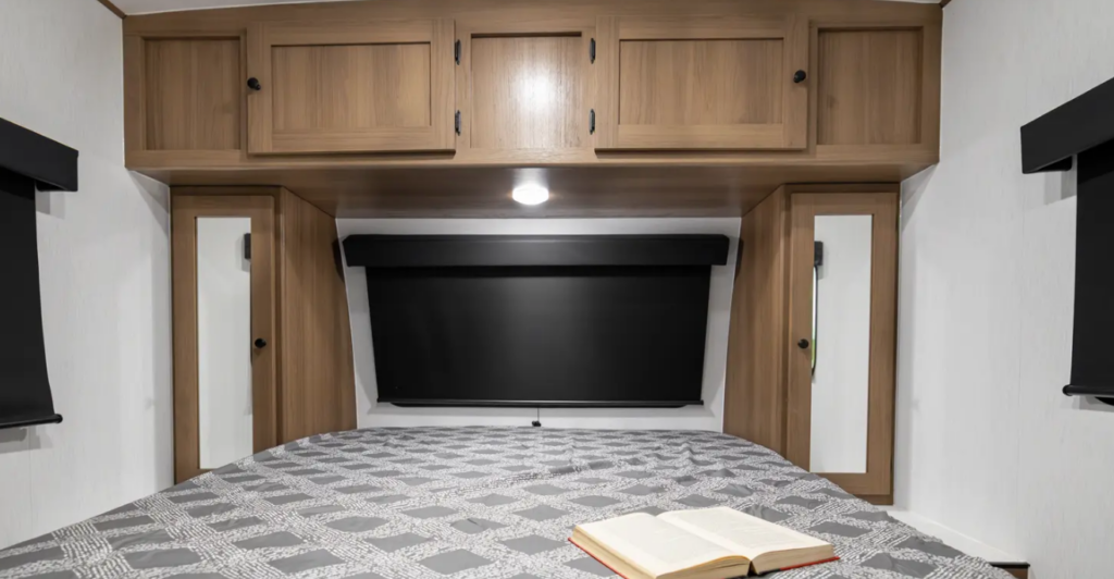 RV bedroom with overhead wood cabinetry, bedside wardrobes with mirrors, blackout window shade, and a bed with an open book on top