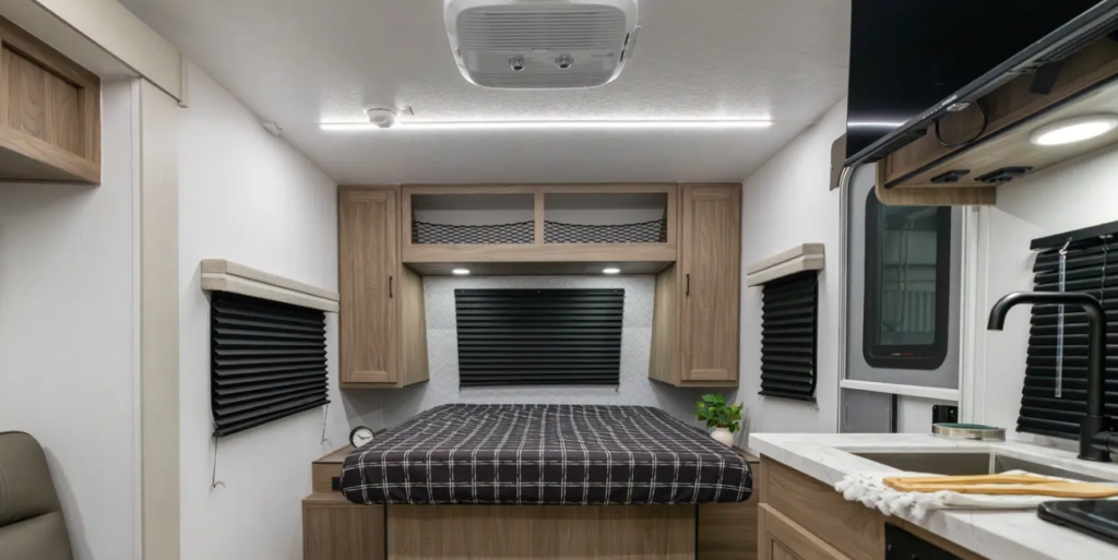 Compact travel trailer interior with rear bed, overhead storage, blackout window shades, kitchenette with sink, and mounted TV.
