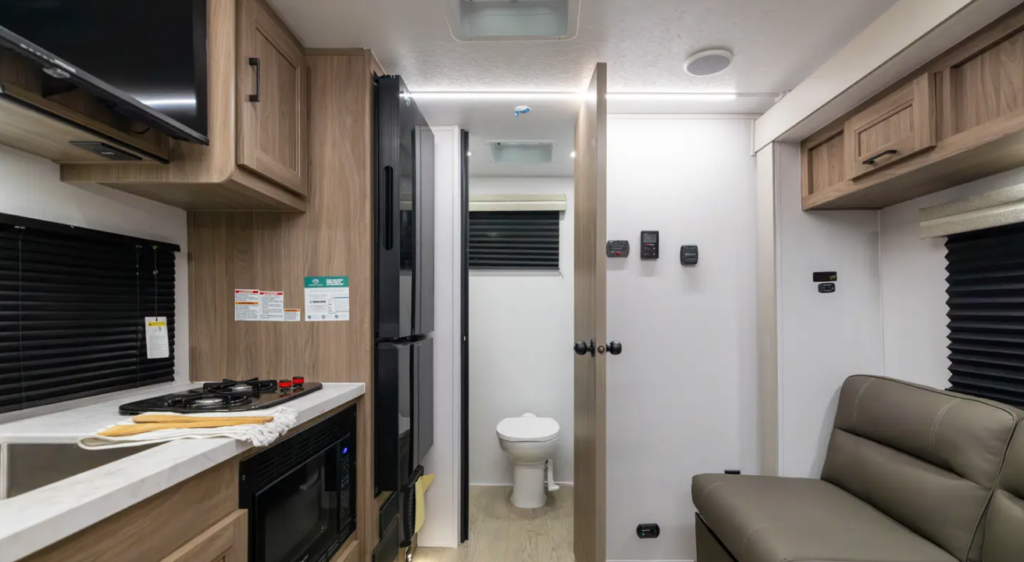 Compact RV interior with kitchenette, refrigerator, leather seating, overhead cabinets, and rear bathroom visible through an open door.