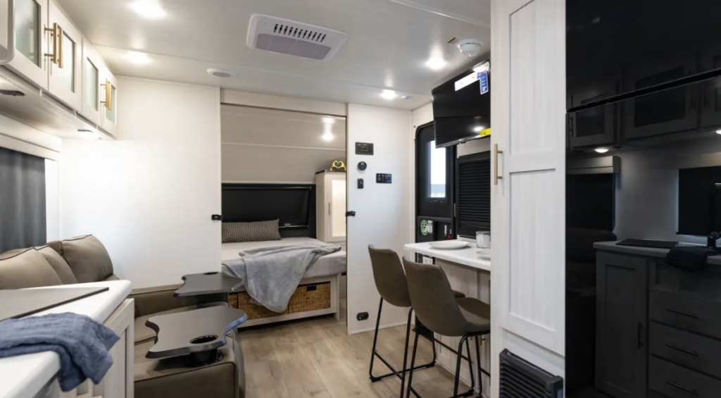Modern RV interior with kitchenette, bar-style dining area, sofa seating, and rear bed with storage baskets underneath.