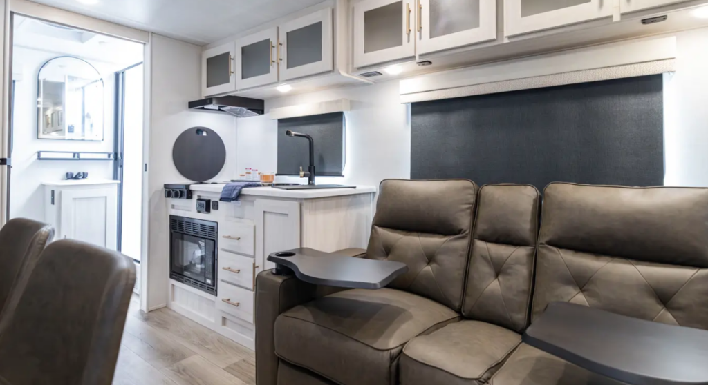 Compact RV interior with reclining theater seating, kitchenette, overhead cabinets, blackout window shades, and bathroom visible in the background