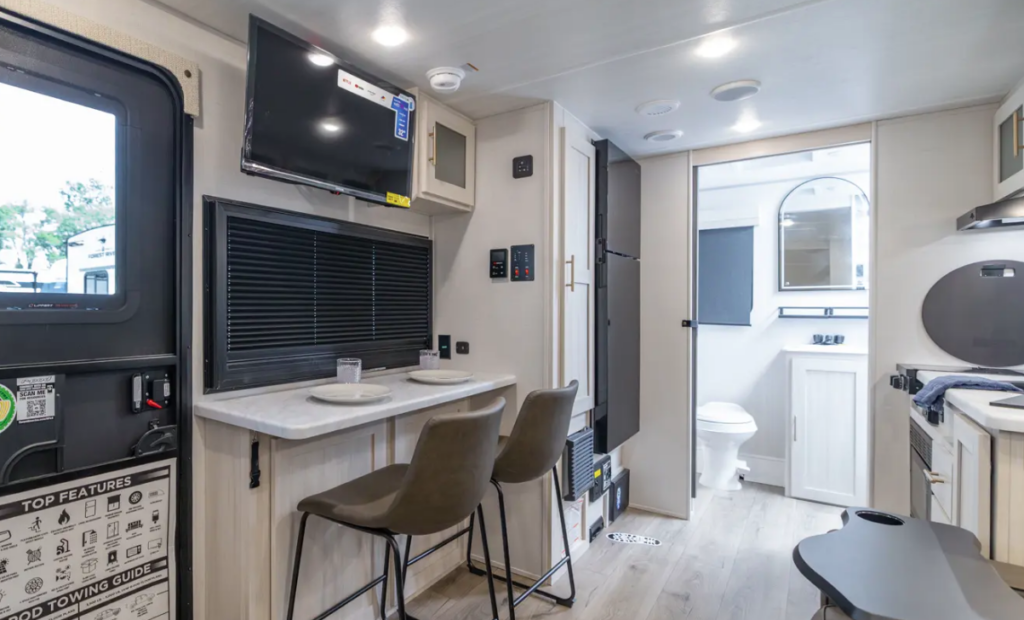 Modern RV interior with bar-style dining area, mounted TV, kitchenette, refrigerator, and rear bathroom visible through an open doorway