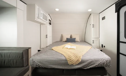 Compact RV bedroom with a large bed, overhead cabinets, accent pillows, and an open book placed on a checkered blanket.