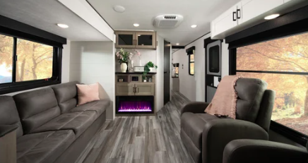Spacious RV living area with sofa seating, recliner chair, large windows, and an entertainment center with an electric fireplace.