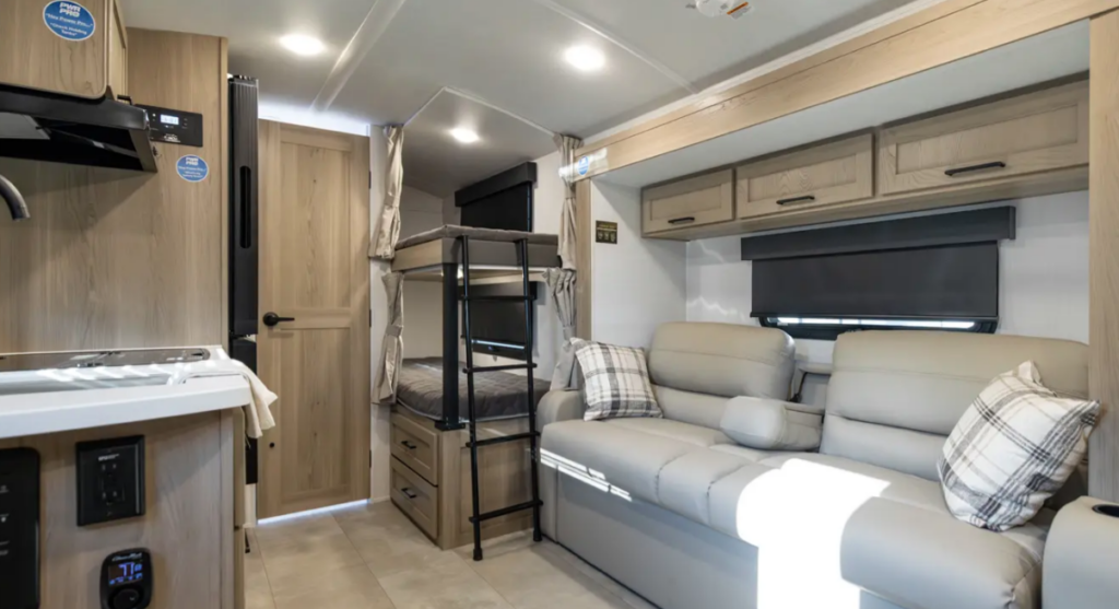 Travel trailer interior with reclining sofa, bunk beds with ladder, overhead storage cabinets, and kitchenette visible in the foreground.