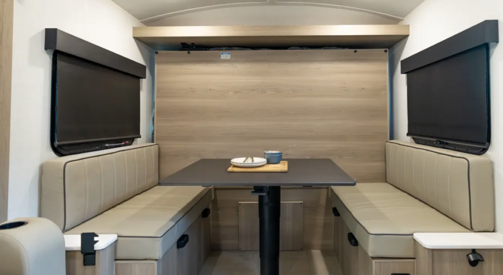 RV dinette area with wraparound bench seating, center table, blackout window shades, and a fold-down bed stored behind the rear wall panel.