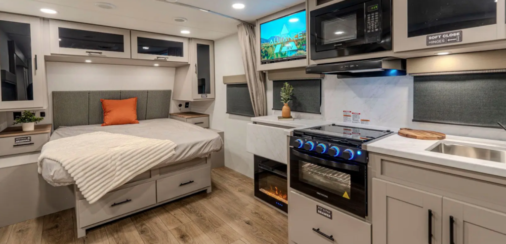 Modern RV interior with rear bed, kitchenette, stove with illuminated knobs, microwave, fireplace, and overhead storage cabinets.