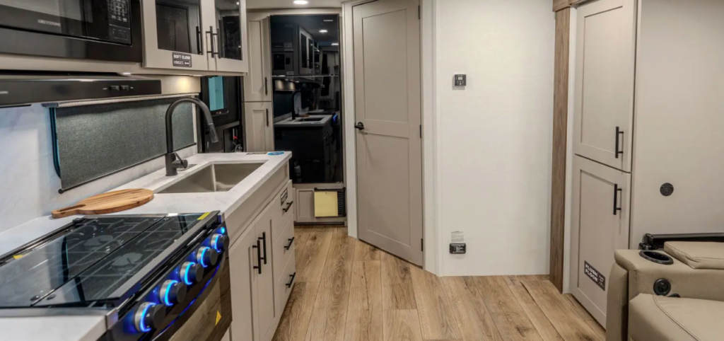 Modern RV interior with kitchenette, stove featuring illuminated knobs, farmhouse-style sink, cabinetry, and hallway leading toward the bathroom and bedroom