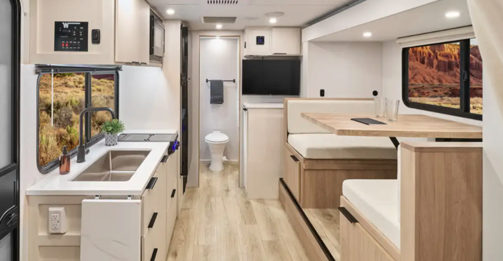 Modern RV interior with kitchenette, booth dinette seating, mounted TV, and rear bathroom visible through the hallway.
