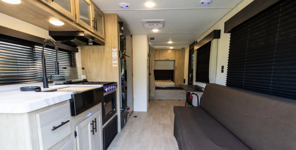 Travel trailer interior with kitchenette, sofa seating, blackout window shades, and rear bedroom visible down the hallway