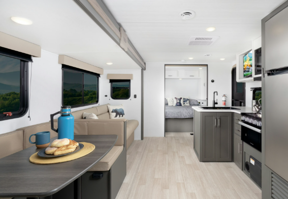 Bright travel trailer interior with booth dinette, sofa seating, full kitchenette, and rear bedroom visible through the hallway