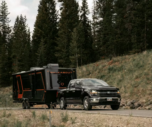 Black pickup truck towing a Keystone Walkabout travel trailer through a forested mountain roadway.
