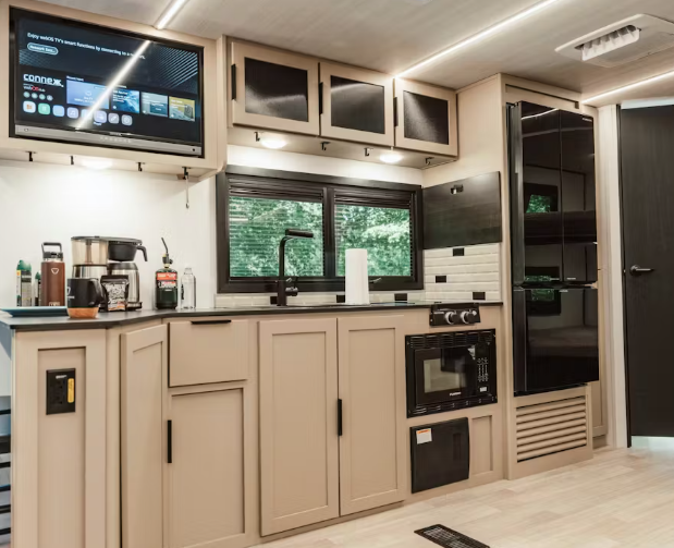 Modern RV kitchenette with farmhouse-style sink, overhead cabinets, smart TV, refrigerator, microwave, and wood-look cabinetry.