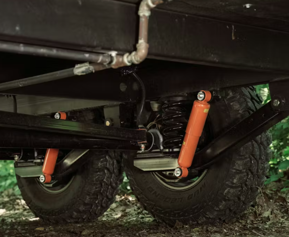 Close-up of an RV independent suspension system with off-road tires, coil springs, and orange shock absorbers underneath the trailer chassis.