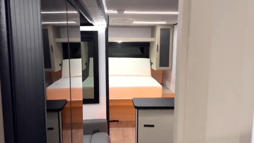 View through an RV hallway showing a fold-down Murphy bed, mirrored wardrobe doors, and compact dining counters on each side.