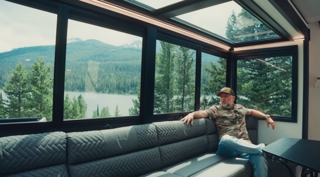 Man relaxing on RV lounge seating beside panoramic windows overlooking a mountain lake and forest landscape