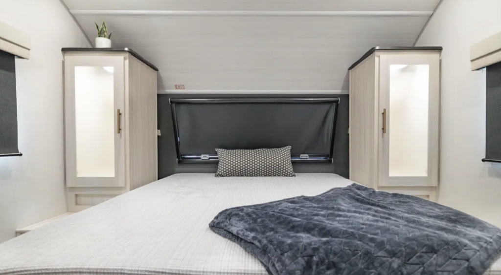 Compact RV bedroom with a large bed, bedside wardrobes, blackout window shade, accent lighting, and decorative bedding
