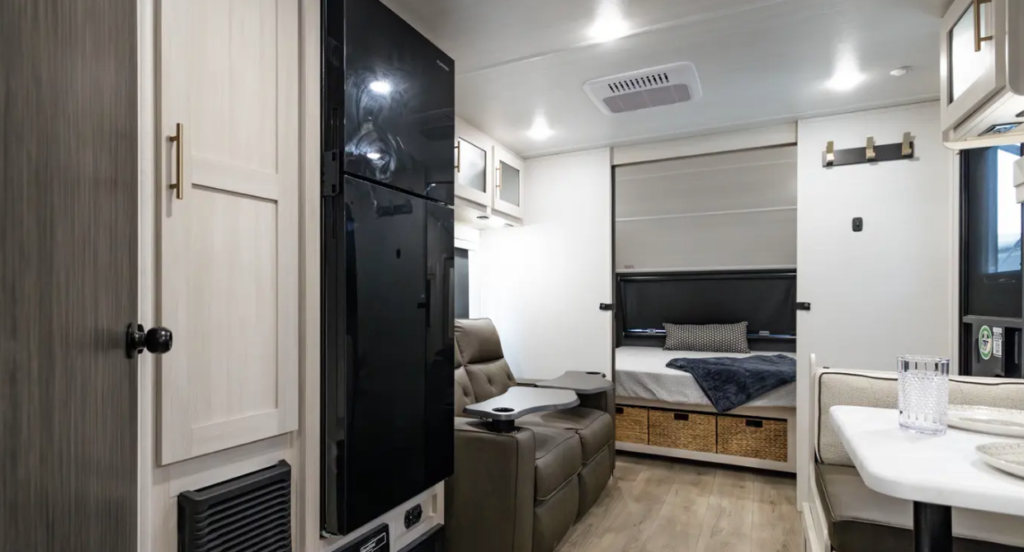 Modern RV interior with booth dinette, recliner seating, tall refrigerator, and rear bed with storage baskets underneath