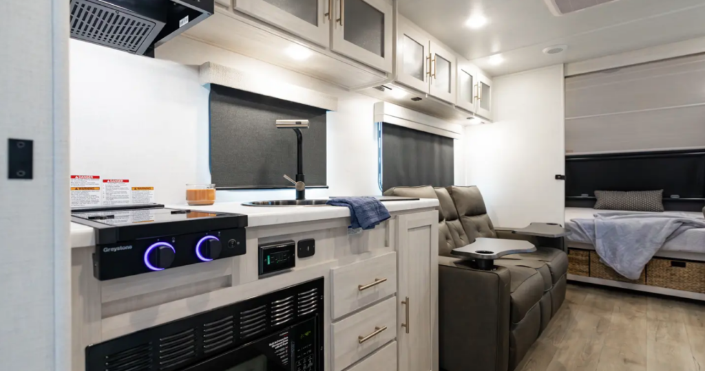 Compact RV interior with kitchenette, illuminated stove controls, recliner seating, overhead cabinets, and rear bed with storage baskets underneath