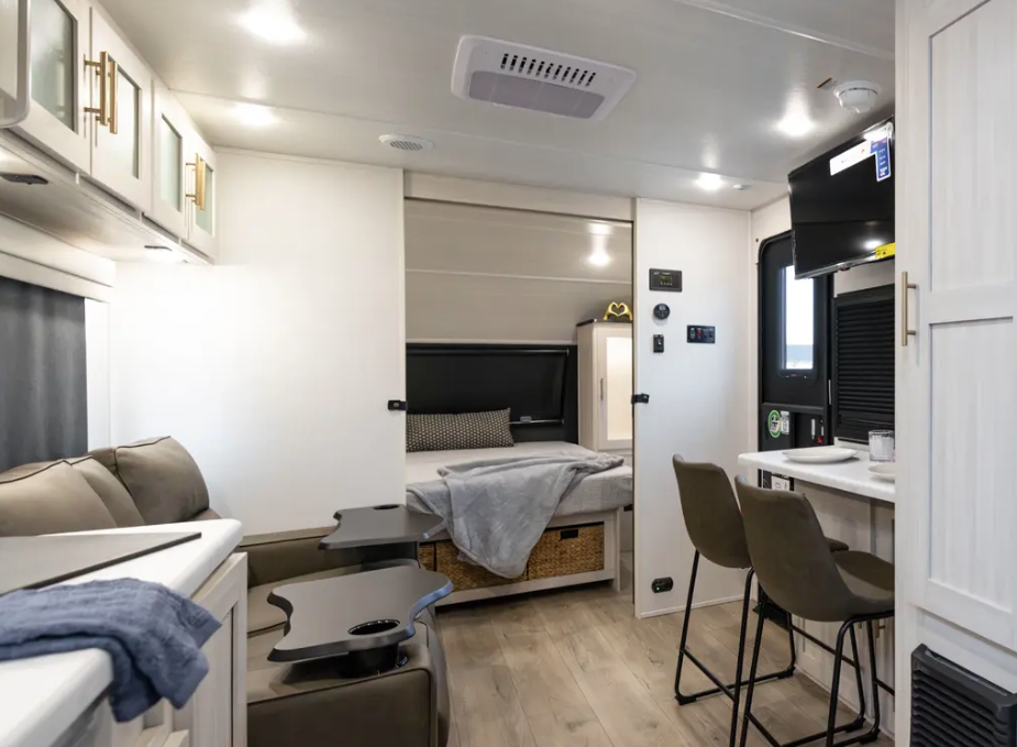 Modern RV interior with kitchenette, recliner seating, bar-style dining area, mounted TV, and rear bed with under-bed storage baskets