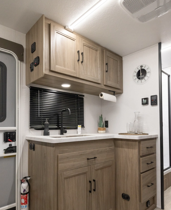 Compact RV kitchenette with wood cabinetry, farmhouse-style sink, countertop workspace, overhead storage, and modern black fixtures