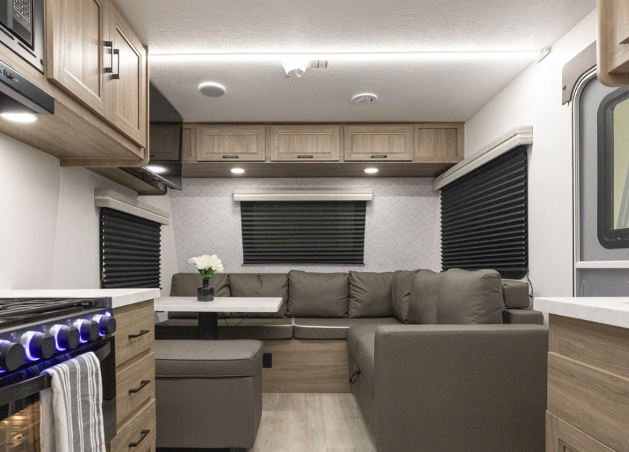 Modern RV living area with sectional sofa seating, dinette table, overhead cabinets, blackout window shades, and kitchenette with illuminated stove controls
