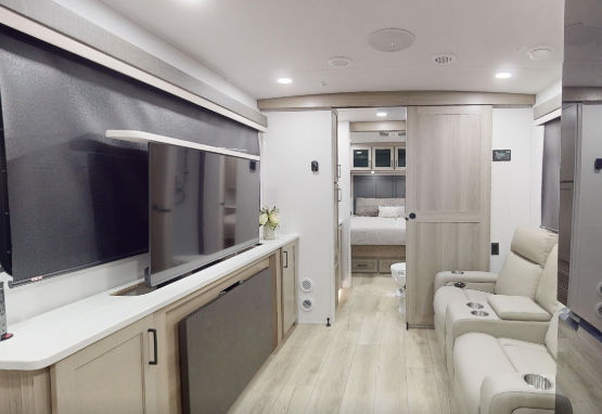 Spacious RV interior with entertainment center, reclining theater seats, sliding bedroom door, and rear sleeping area visible through the hallway