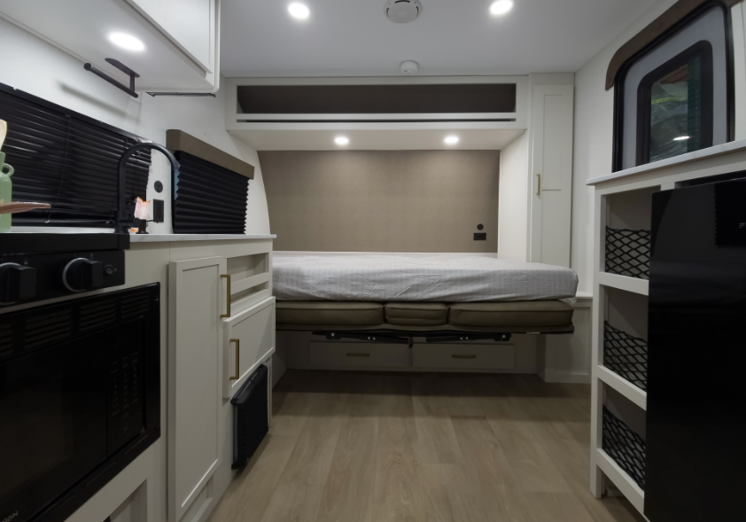 Compact RV interior with kitchenette, storage shelving, mini refrigerator, and Murphy bed lowered into sleeping position