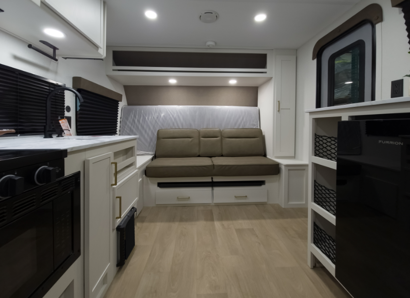 Compact RV interior with kitchenette, storage shelving, mini refrigerator, and convertible sofa positioned in front of a folded Murphy bed
