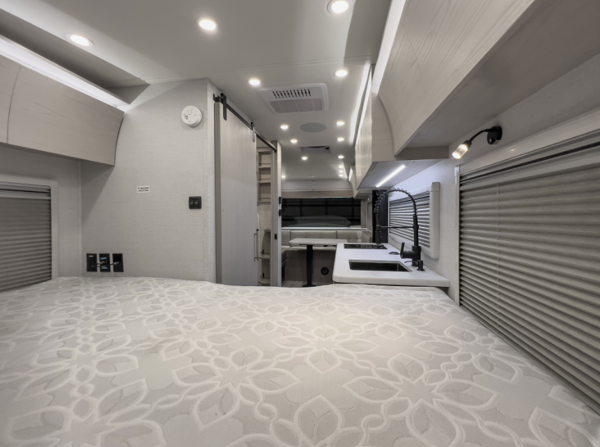 Modern RV interior viewed from the bed area, featuring a compact kitchenette, overhead lighting, sliding bathroom door, and rear dinette seating