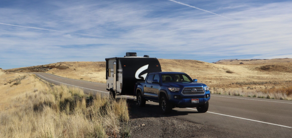 Blue pickup truck towing a compact travel trailer along a scenic desert highway with rolling hills and open skies in the background.