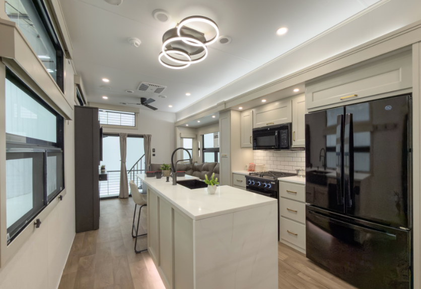 Luxury RV kitchen with a large island, residential refrigerator, gas range, modern lighting fixture, and upscale white cabinetry with gold accents