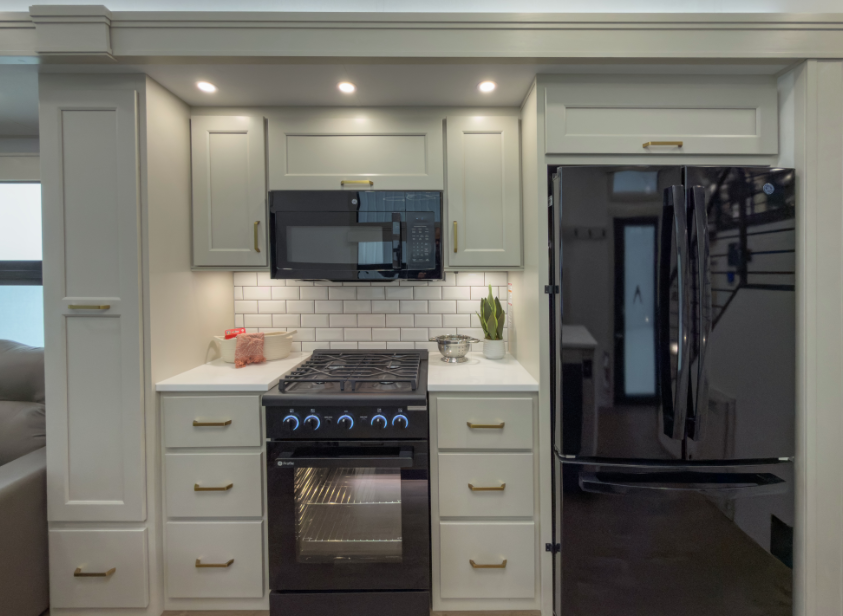Residential-style RV kitchen with full-size refrigerator, gas range and oven, microwave, subway tile backsplash, and white cabinetry with gold hardware