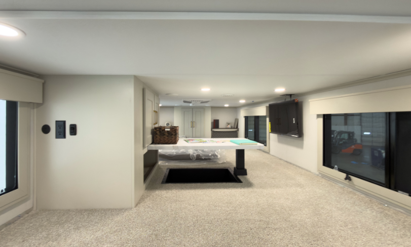 Spacious loft area inside a luxury RV featuring carpeted flooring, built-in entertainment center, low-profile ceiling, and lounge space with table and mattress setup