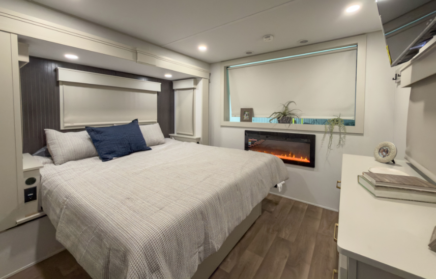 Luxury RV bedroom with a queen bed, built-in fireplace beneath a large window, modern cabinetry, and soft ambient lighting