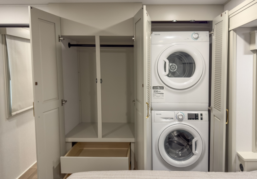 Luxury RV bedroom storage area featuring a wardrobe closet with drawers beside a stacked washer and dryer unit hidden behind cabinet doors