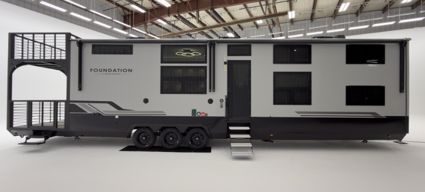 Large Foundation luxury travel trailer with triple axles, modern gray-and-white exterior, rooftop deck platform, and multiple large windows displayed indoors.