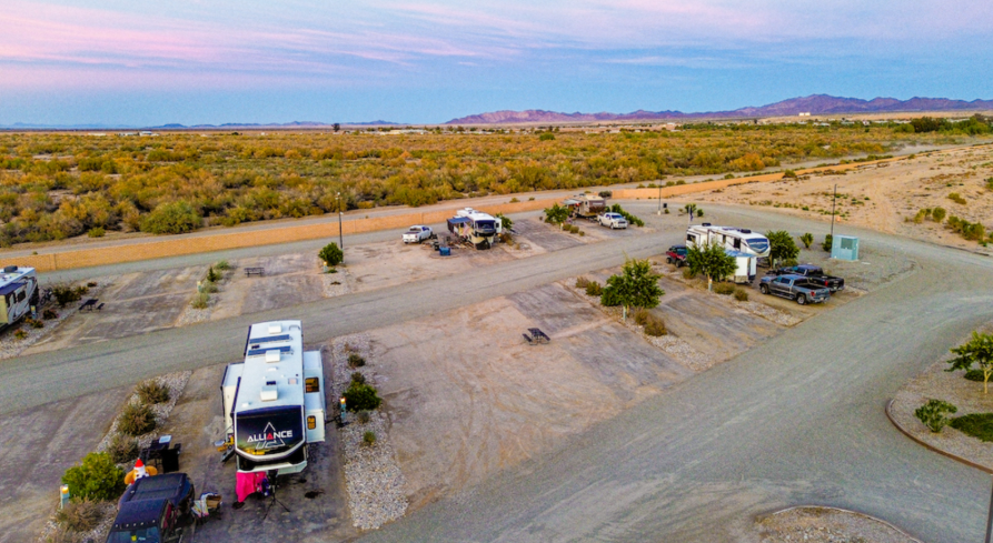 Aerial view of an RV campground in a desert landscape with scattered campsites, parked RVs, gravel roads, and distant mountain ranges at sunset