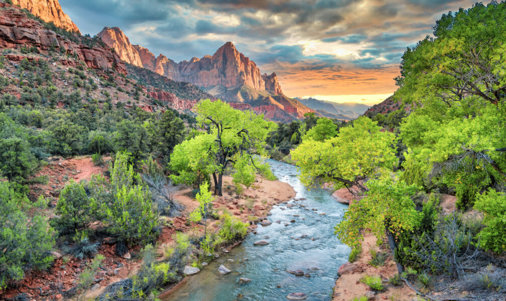 Scenic desert canyon landscape with a flowing river, lush green trees, red rock cliffs, and dramatic sunset clouds over towering mountain formations