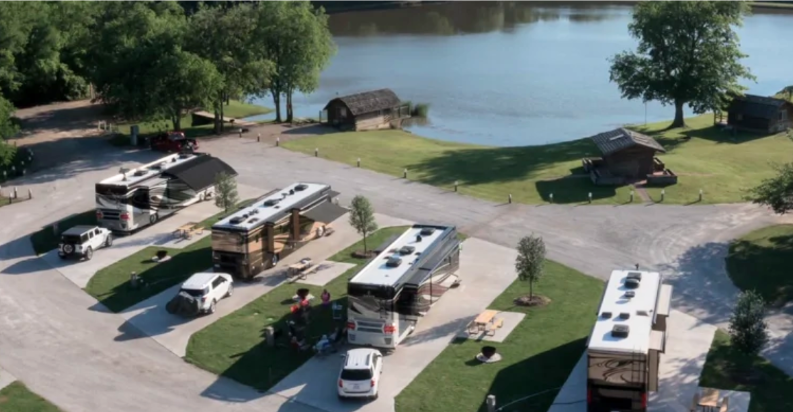 Aerial view of a lakeside RV resort with spacious concrete campsites, luxury motorhomes, picnic areas, and waterfront cabins surrounded by trees.