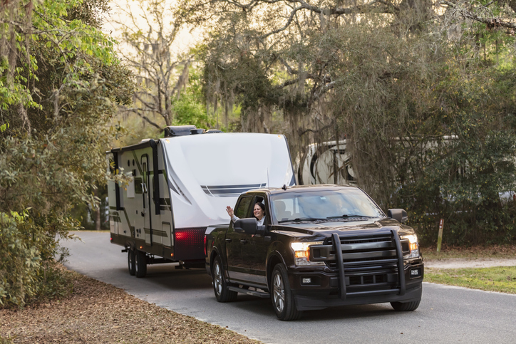 A camper trailer on a rural road. A mature multiracial couple is inside the pick-up truck. The woman is waving out the window at the camera. The man is driving.